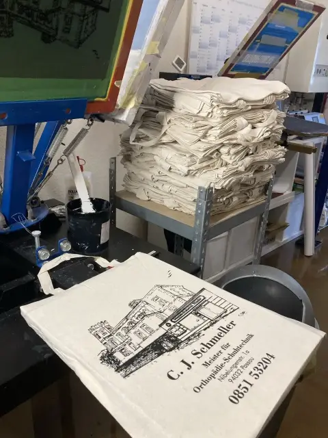 500 im Siebdruck bedruckte Baumwolltaschen mit Fassaden-Logo von C. J. Schmeller Orthopädie-Schuhtechnik Passau vor Druckkarussell