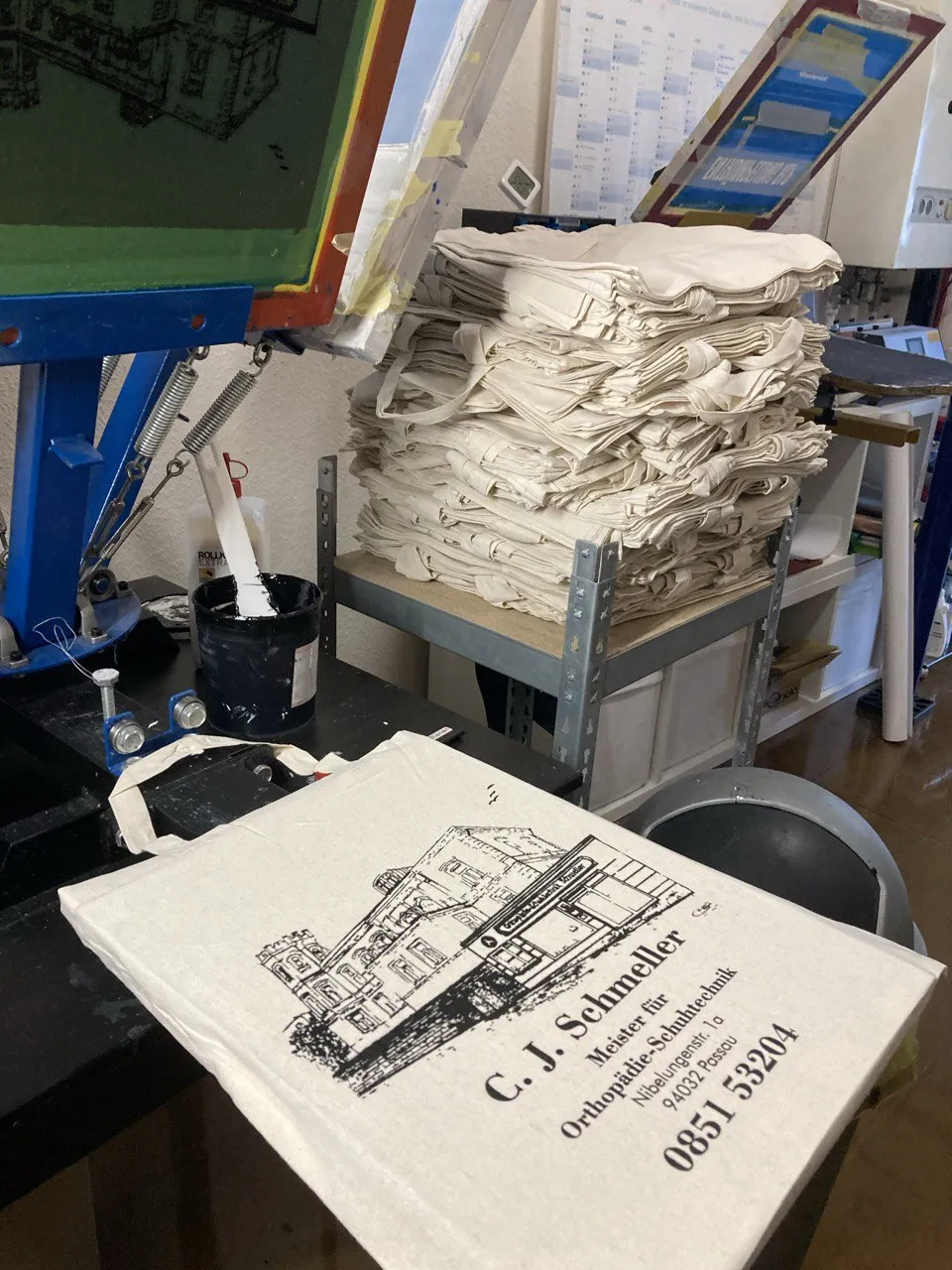 500 im Siebdruck bedruckte Baumwolltaschen mit Fassaden-Logo von C. J. Schmeller Orthopädie-Schuhtechnik Passau vor Druckkarussell